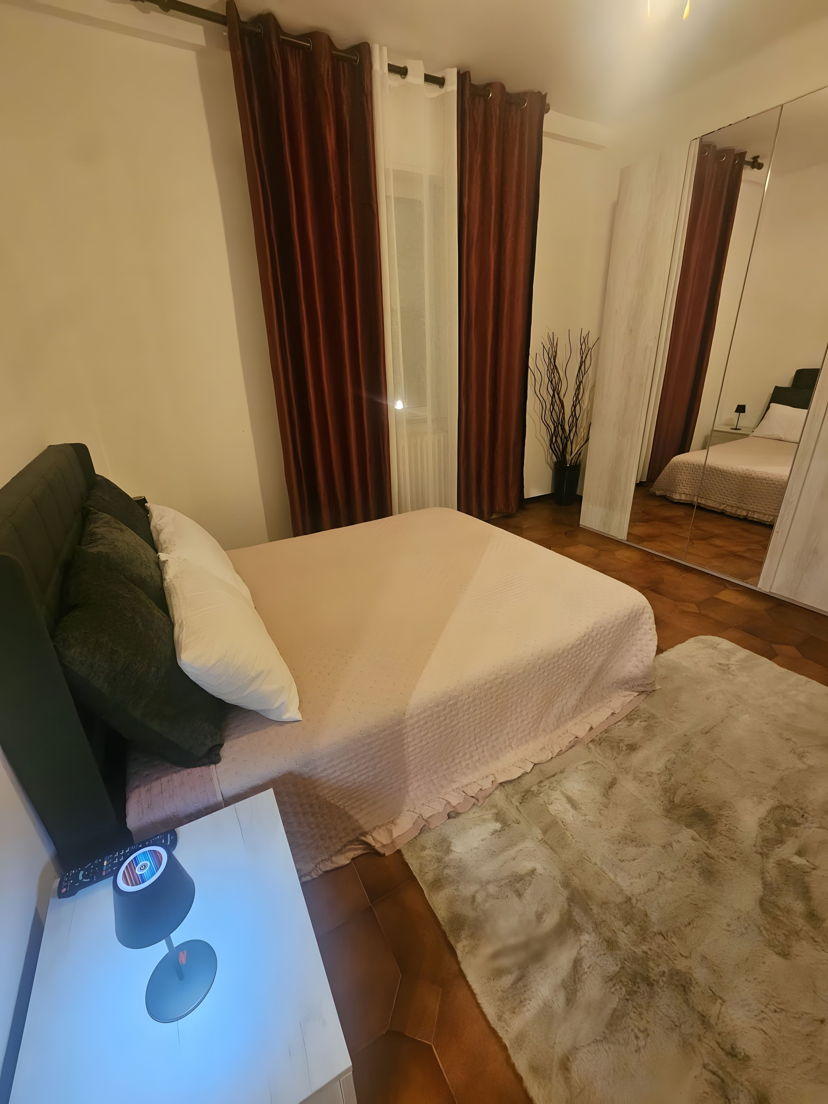 Ein Schlafzimmer mit einem Bett, einem Nachttisch mit Lampe, einem Spiegel und einem Fenster mit Vorhängen.