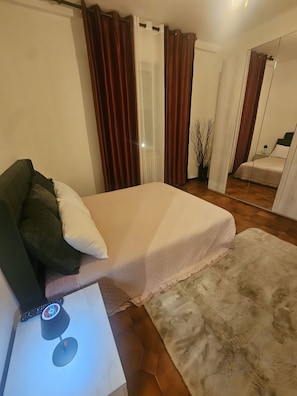 Une chambre à coucher avec un lit, une table de chevet avec une lampe, un miroir et une fenêtre avec des rideaux.