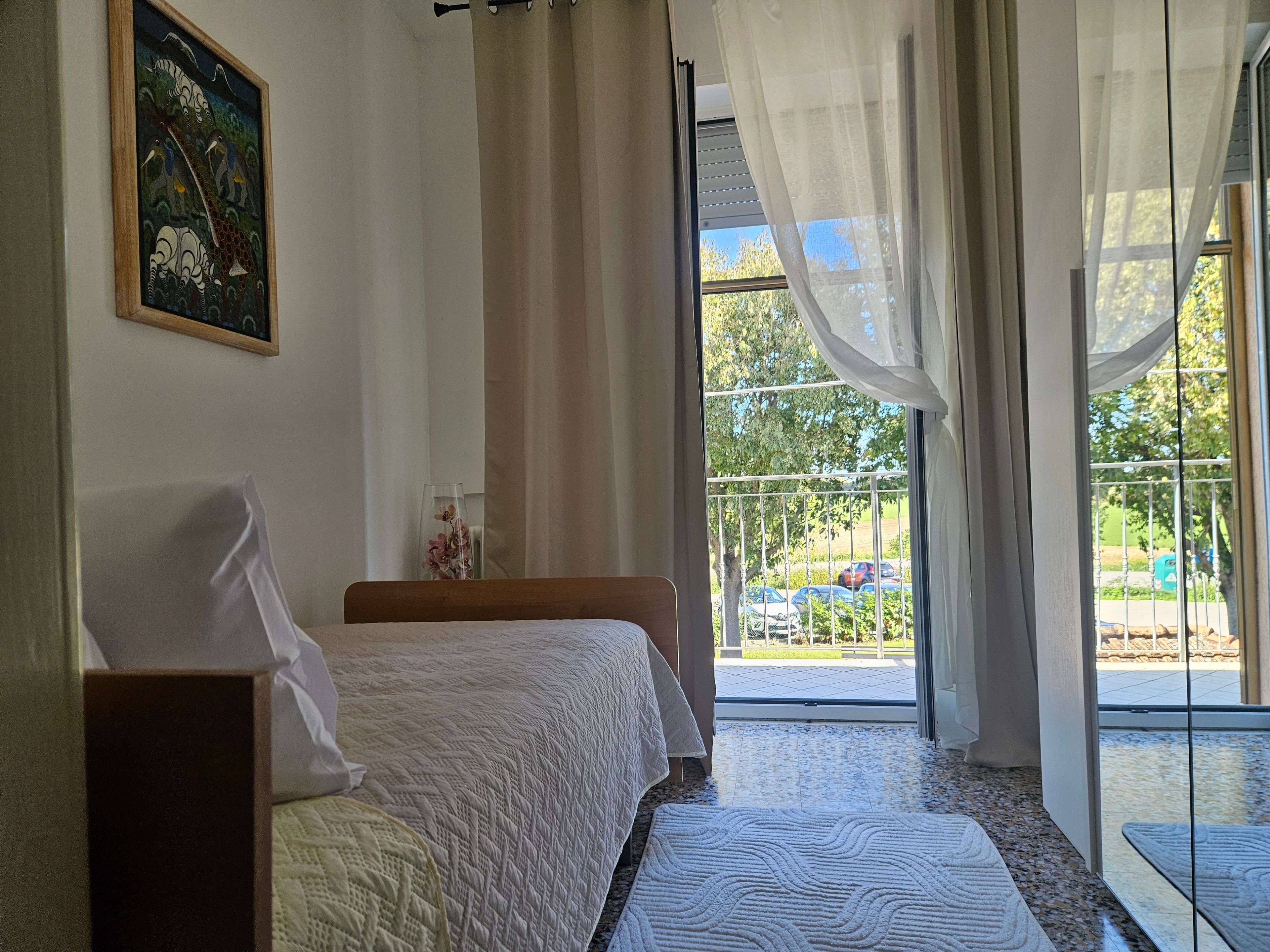 Ein Schlafzimmer mit zwei Betten, einem Fenster mit Blick auf geparkte Autos und Grünflächen, sowie einem Bild an der Wand.