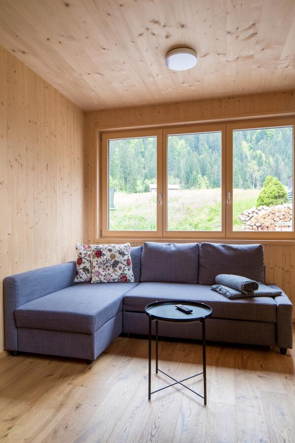 Ein Holzinnenraum mit einem Ecksofa, einem kleinen runden Tisch und einem großen Fenster mit Blick auf Grünflächen.