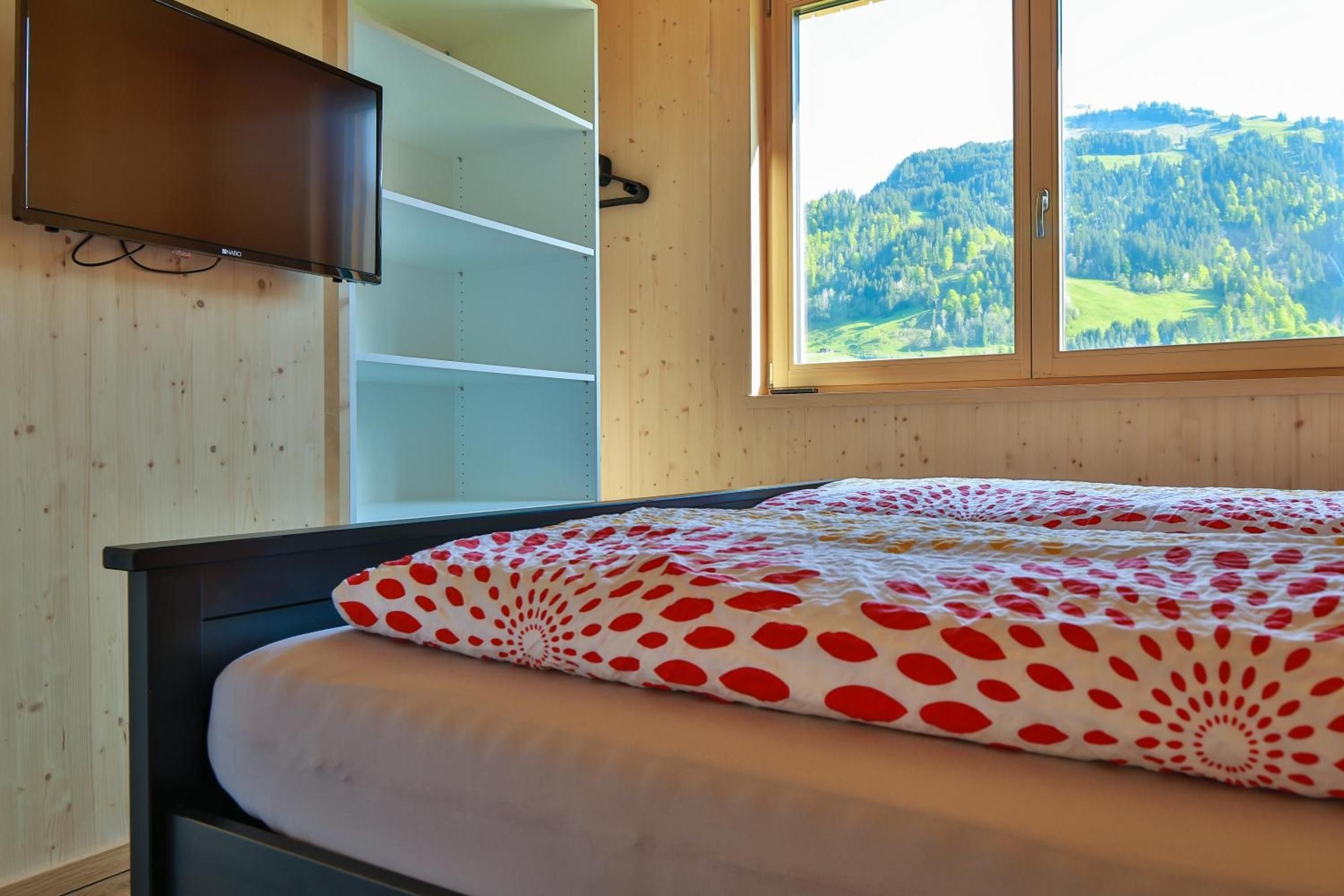 Ein Schlafzimmer mit einem Bett, einem an der Wand befestigten Fernseher und einem Fenster mit Blick auf eine bewaldete Landschaft.