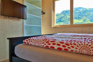 Een slaapkamer met een bed, een aan de muur bevestigde televisie en een raam met uitzicht op een bebost landschap.