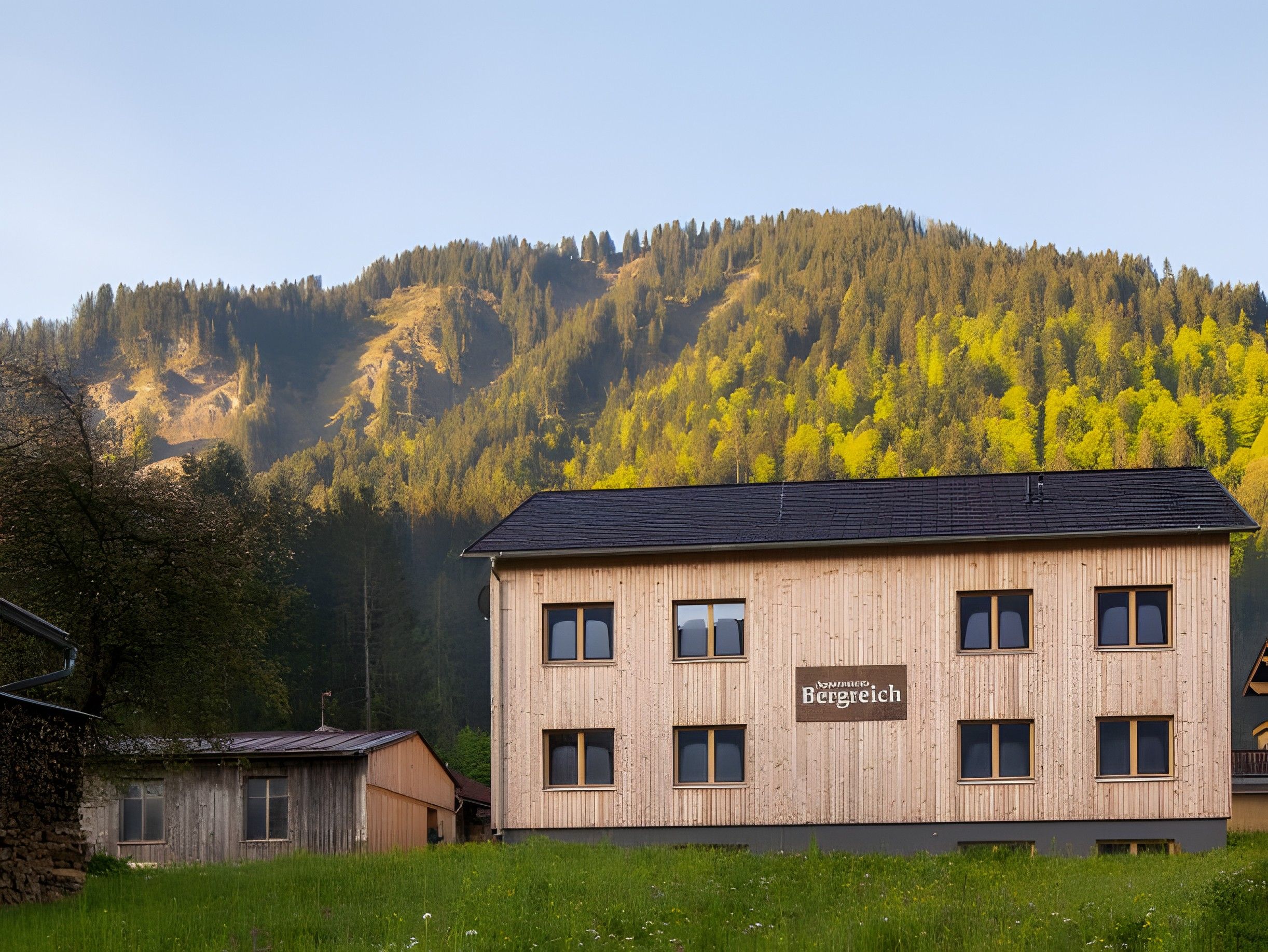 Ein zweistöckiges Holzhaus mit dem Schild 'Bergreich' an der Seite, vor einer bewaldeten Berglandschaft.