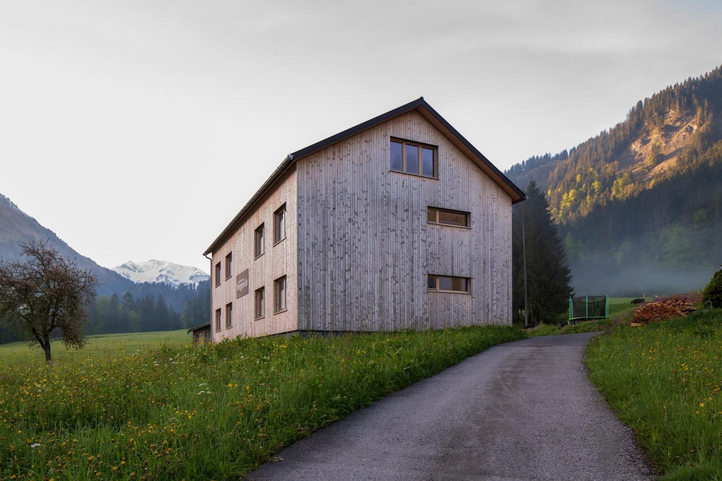 Ein Holzhaus mit geneigtem Dach, einer gepflasterten Auffahrt und einer Bergkulisse im Hintergrund.