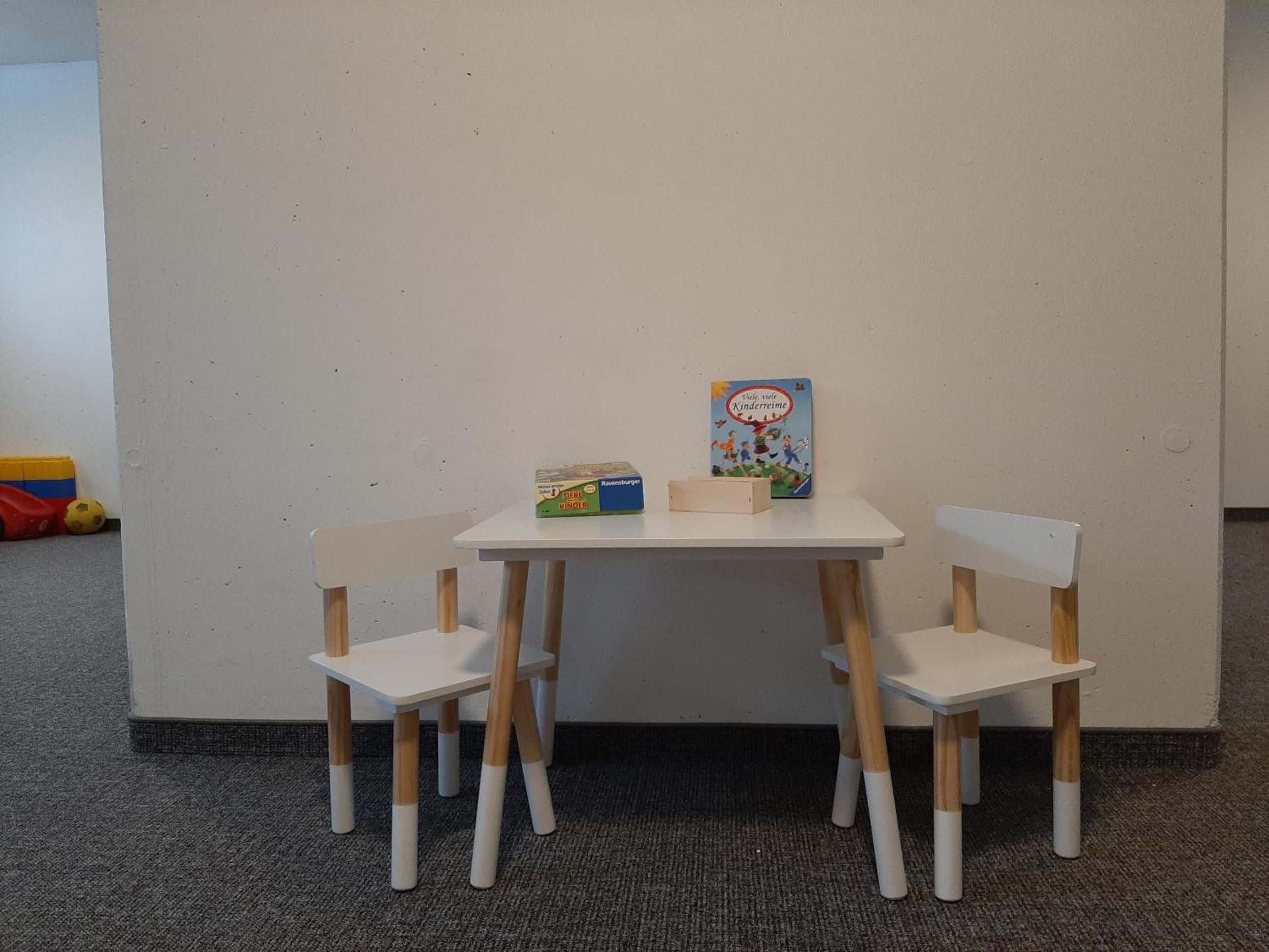 Ein kleiner weißer Tisch mit zwei hölzernen Stühlen, einem Kinderbuch und einem Spielzeugauto.