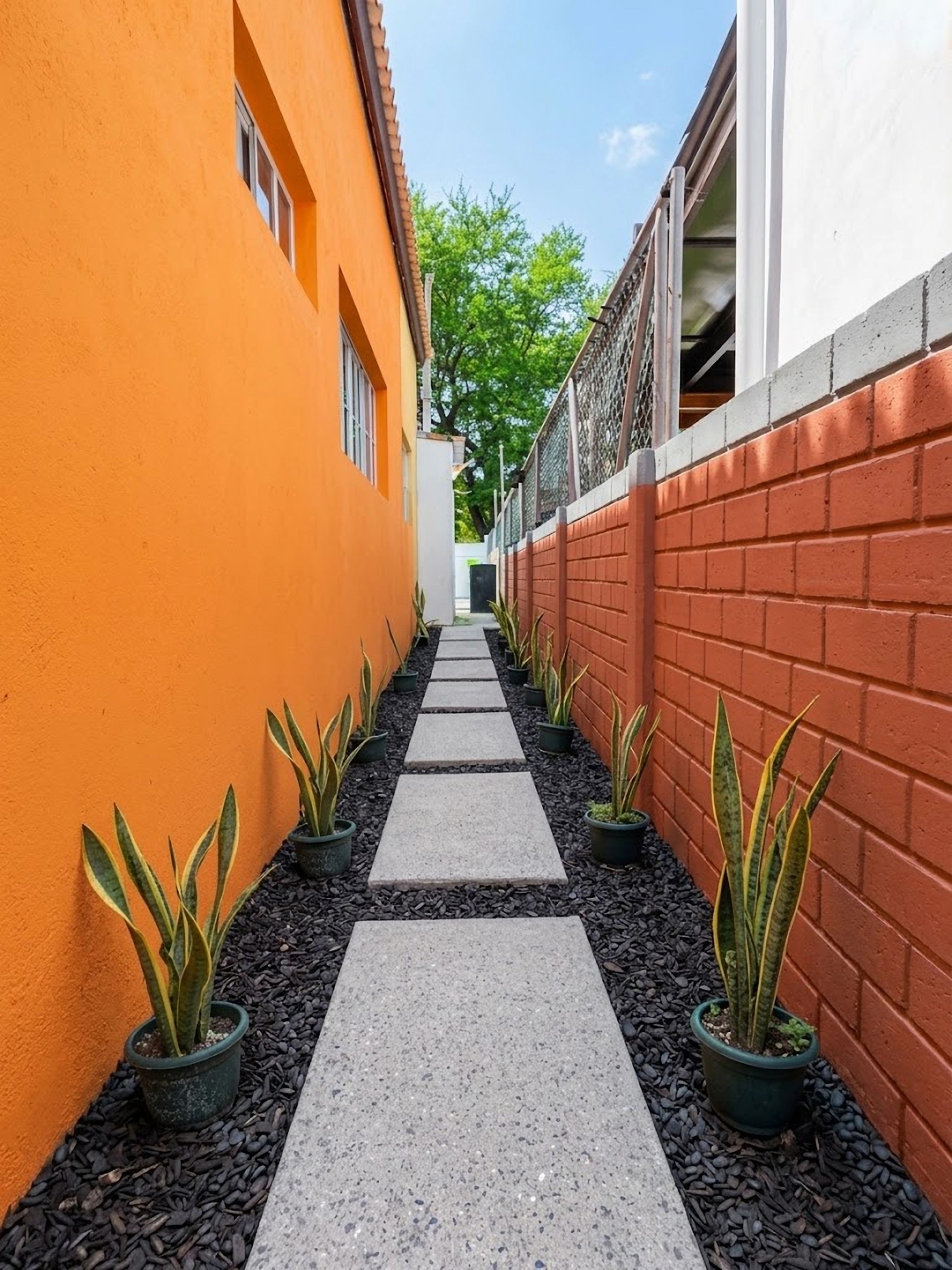 Un étroit sentier bordé de bâtiments, avec un mur de briques d’un côté et un mur orange de l’autre, qui mène à une porte.
