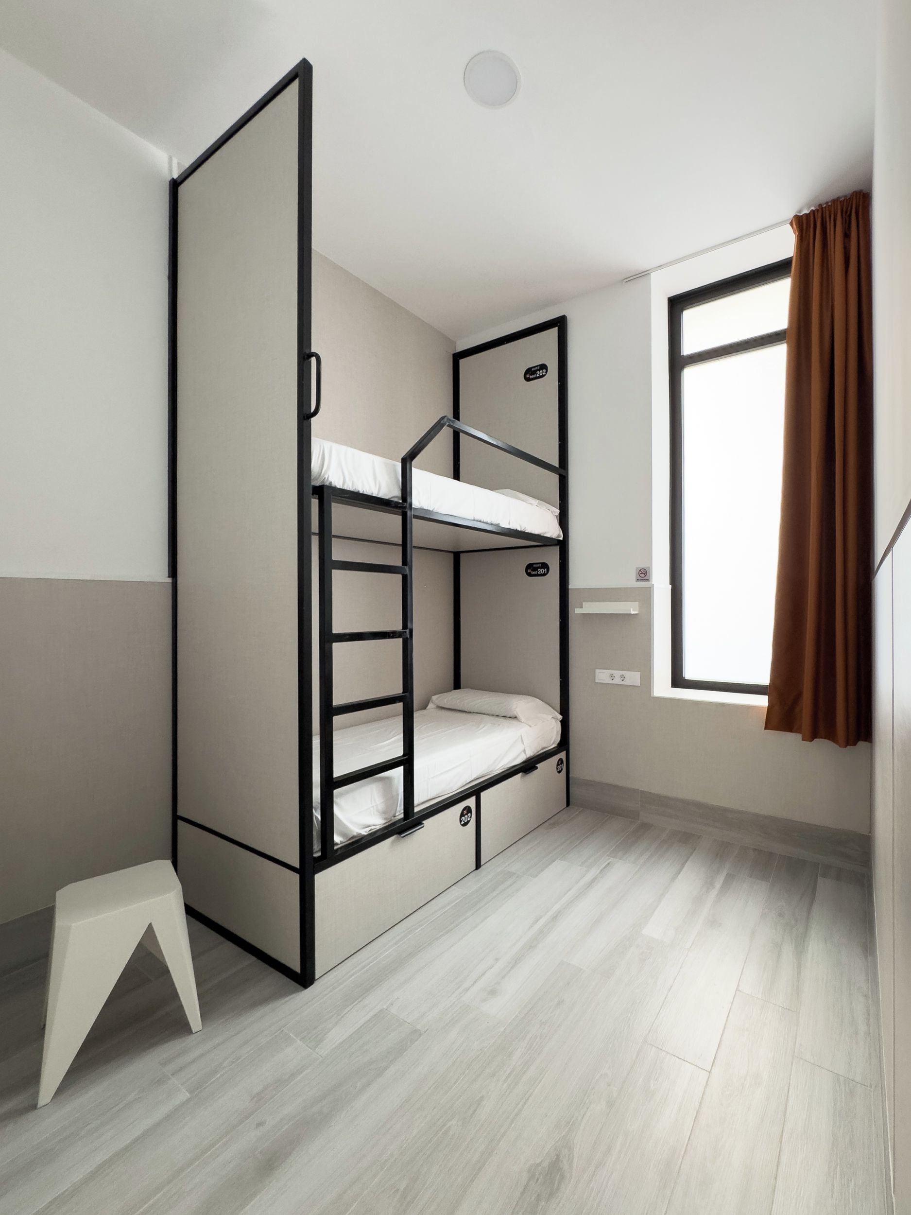 Un lit superposé avec une cloison pliable, un petit tabouret blanc et une fenêtre avec des rideaux marron.