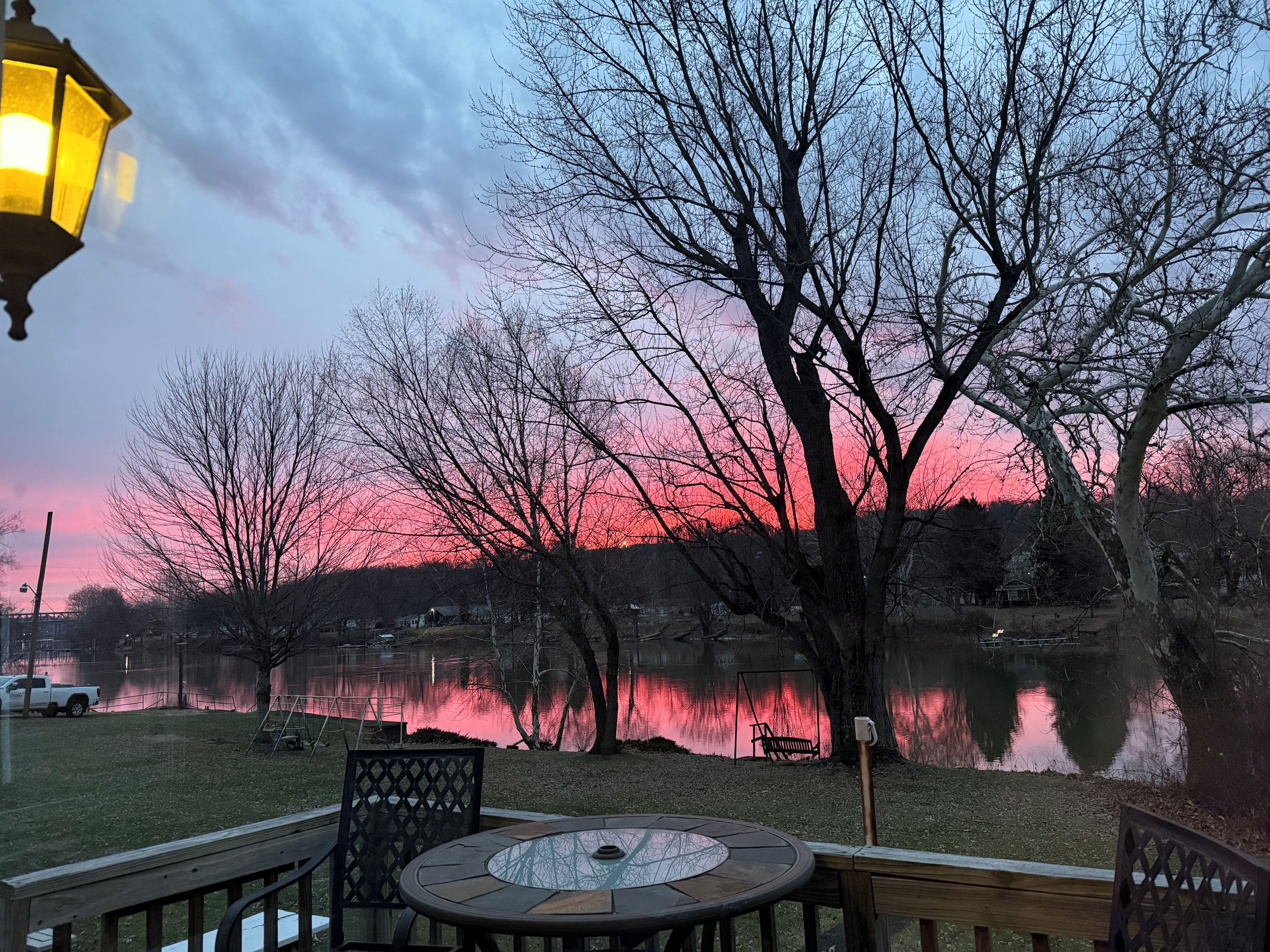 A lakeside patio with a table and chairs, surrounded by trees and a view of a sunset over the water.