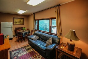 A cozy living room with a leather sofa, a dining table, a refrigerator, and a window with curtains.