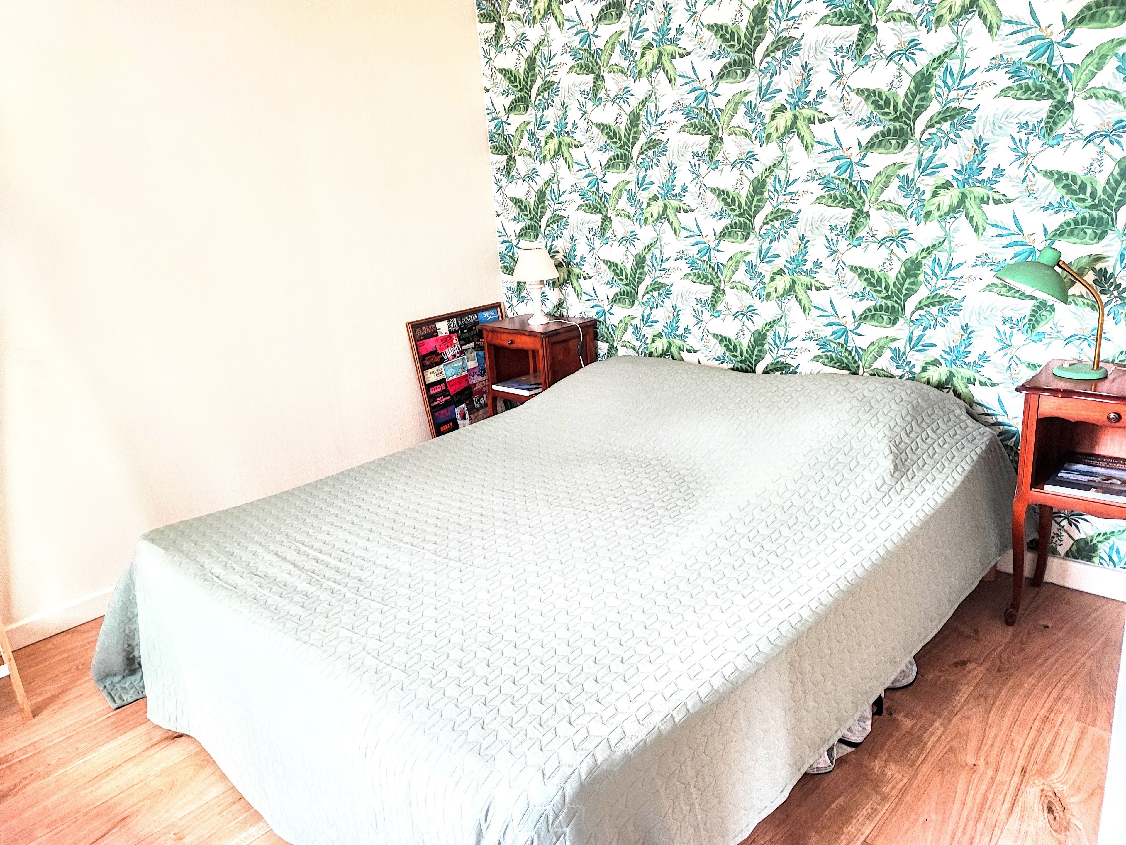 Una cama bien hecha con un edredón texturizado, una mesita de noche de madera con una lámpara y una estantería contra una pared con estampado de hojas tropicales.