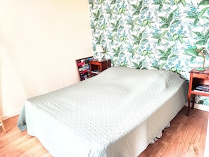 A neatly made bed with a textured blanket, a wooden nightstand with a lamp, and a bookshelf against a wall with a tropical leaf pattern. - Apartment 'Studio Cozy à pied de la Plage' with Wi-Fi (Guilvinec)
