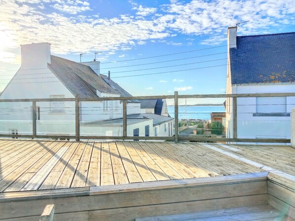 Une terrasse en bois dotée d’une balustrade en verre, offrant une vue sur une zone côtière avec des bâtiments et un ciel dégagé.