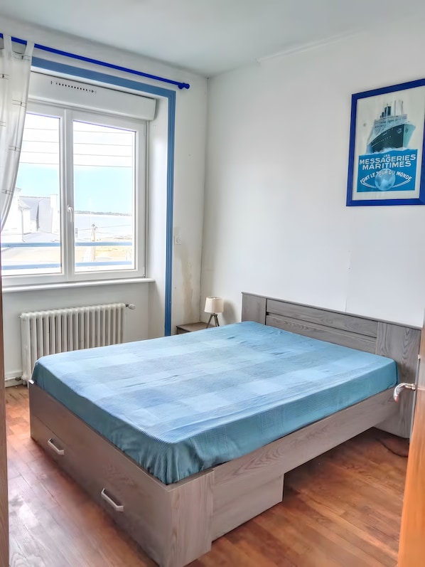 A bedroom with a bed, a window with curtains, a radiator, a lamp, and a framed picture on the wall.