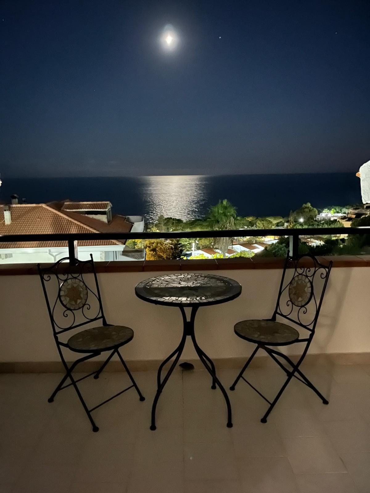 Een balkon met twee stoelen en een kleine tafel, met uitzicht op een kustgebied met gebouwen en zee.