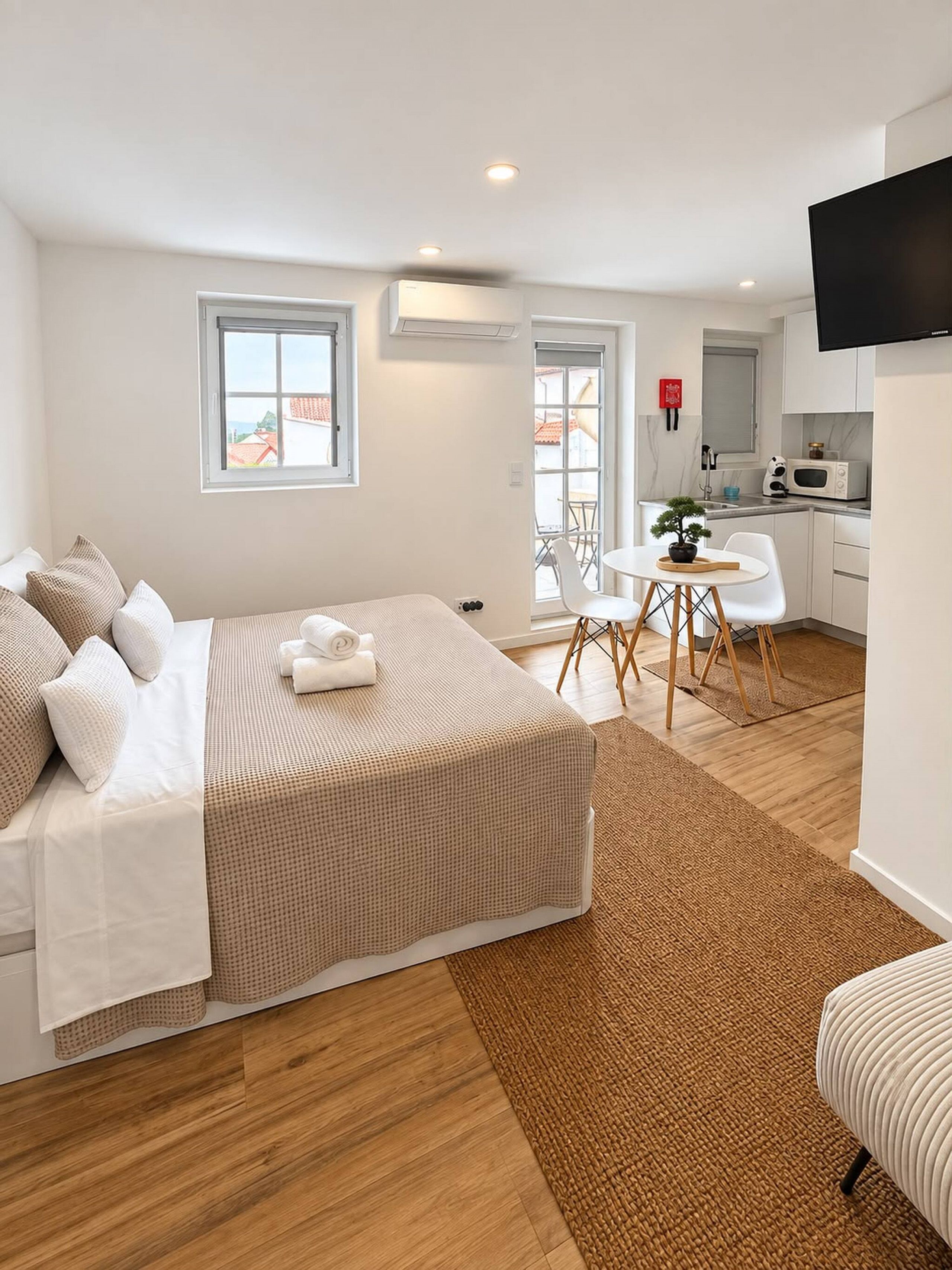 A bedroom with a bed, a small dining area, a kitchen, and a TV mounted on the wall.