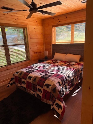 Un dormitorio de cabaña de madera con una cama, un ventilador de techo y ventanas con persianas.