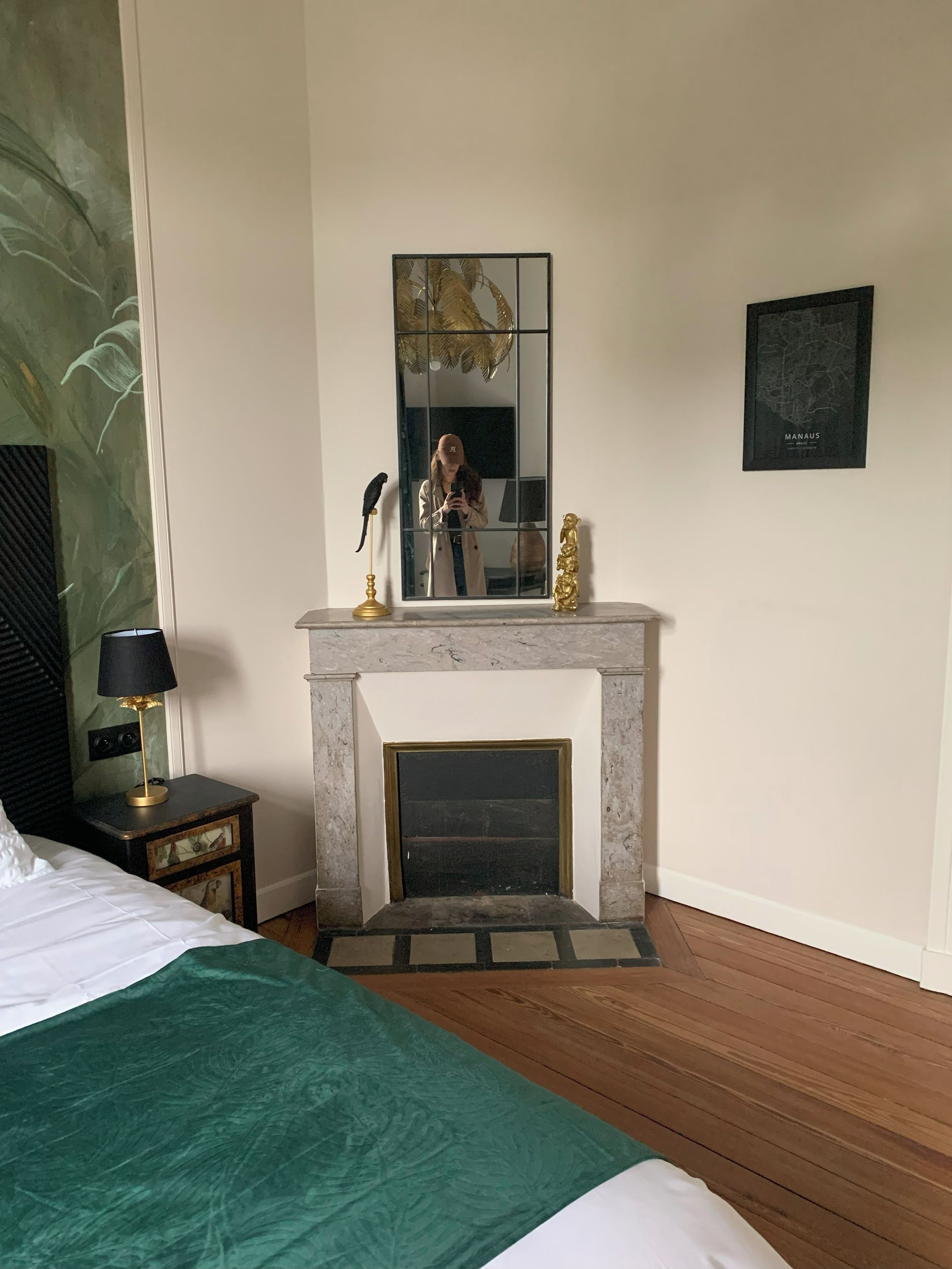 Un dormitorio con cama, una mesita de noche con lámpara, una chimenea y un espejo que refleja a una persona.