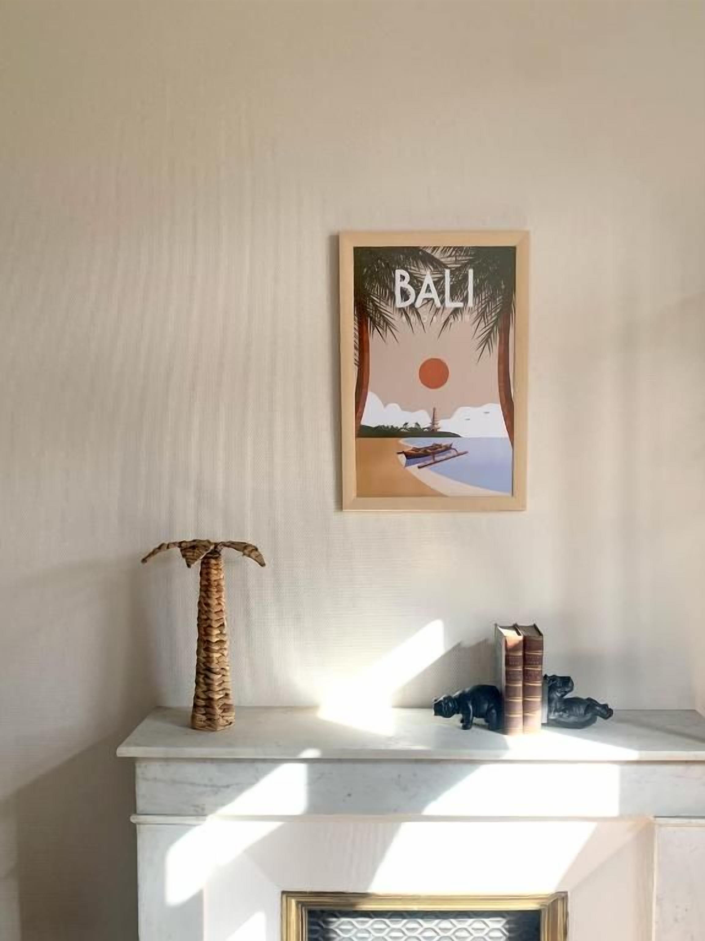 Una obra enmarcada de Bali en una pared, una planta tejida sobre una repisa y una pequeña figurita.