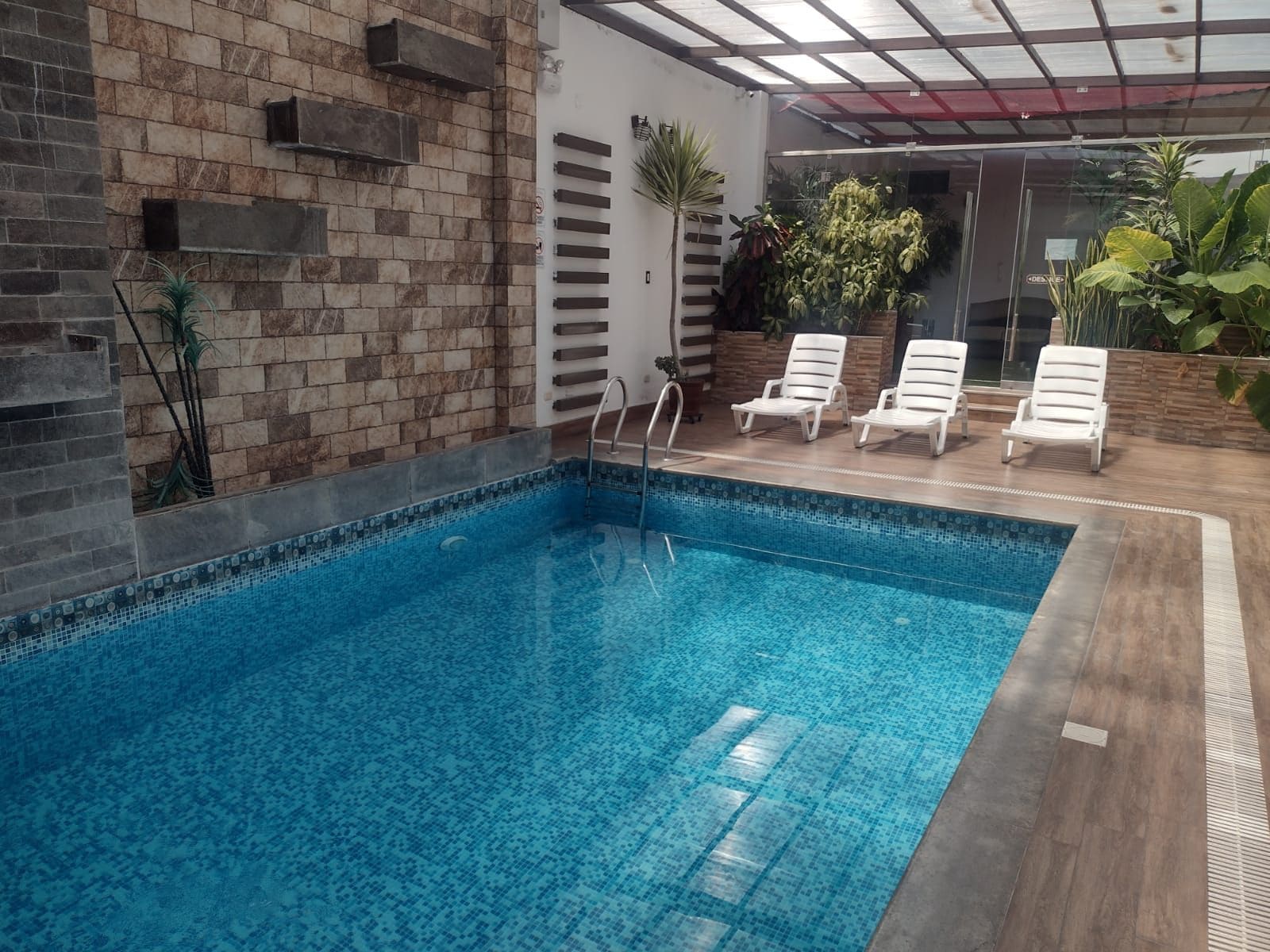 Una piscina interior con borde de azulejos, una escalera de metal y sillones blancos para tomar el sol.