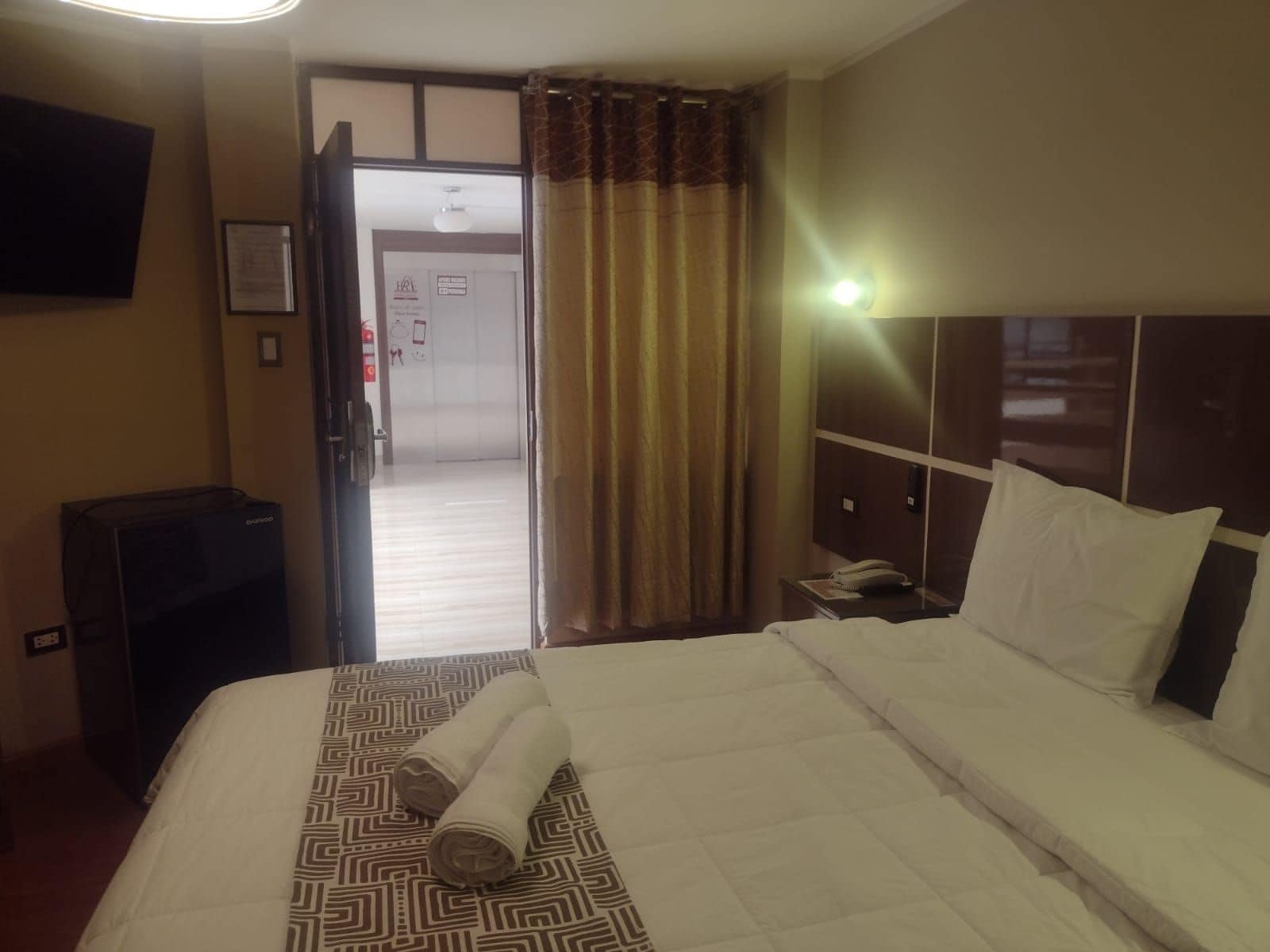 Una habitación de hotel con una cama, una mesita de noche con un teléfono, un televisor montado en la pared y una puerta que da al pasillo.