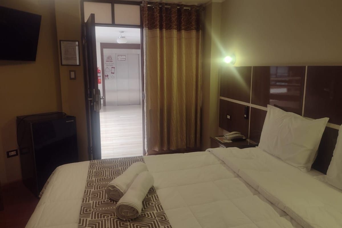 Habitació d'hotel amb un llit, una taula de nit amb un telèfon, una televisió penjada a la paret i una porta que dóna al passadís.
