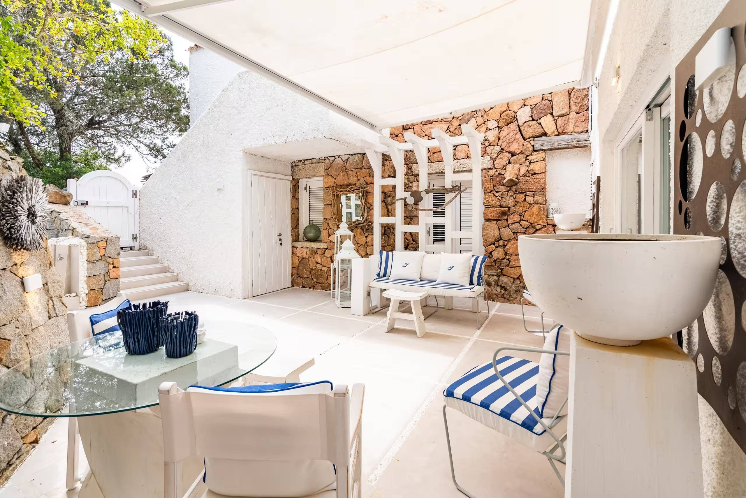 Patio esterno soleggiato con muro in pietra, mobili bianchi e tavolo in vetro.