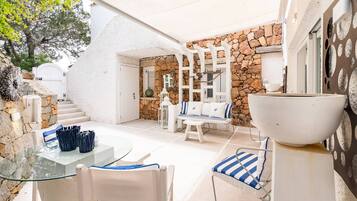 Patio esterno soleggiato con muro in pietra, mobili bianchi e tavolo in vetro.