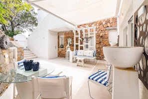 Patio esterno soleggiato con muro in pietra, mobili bianchi e tavolo in vetro.