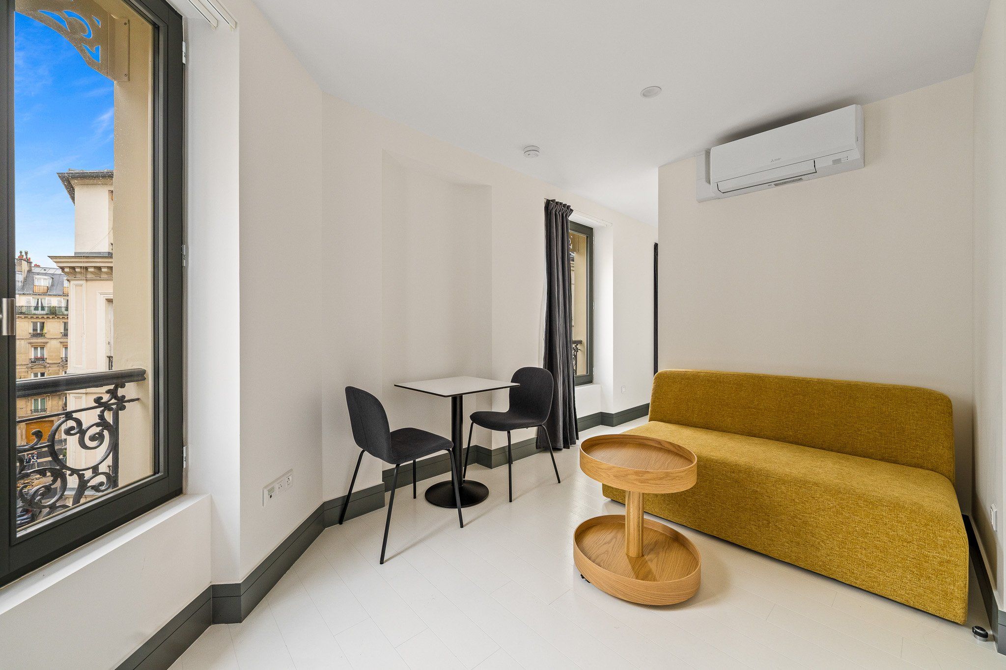 A small, modern room with a sofa, two chairs, a small table, and a window with a view of buildings.