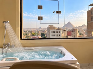 A bathroom with a bathtub, shower, and a window with a view of a pyramid. - Wateen Pyramids View (Giza)
