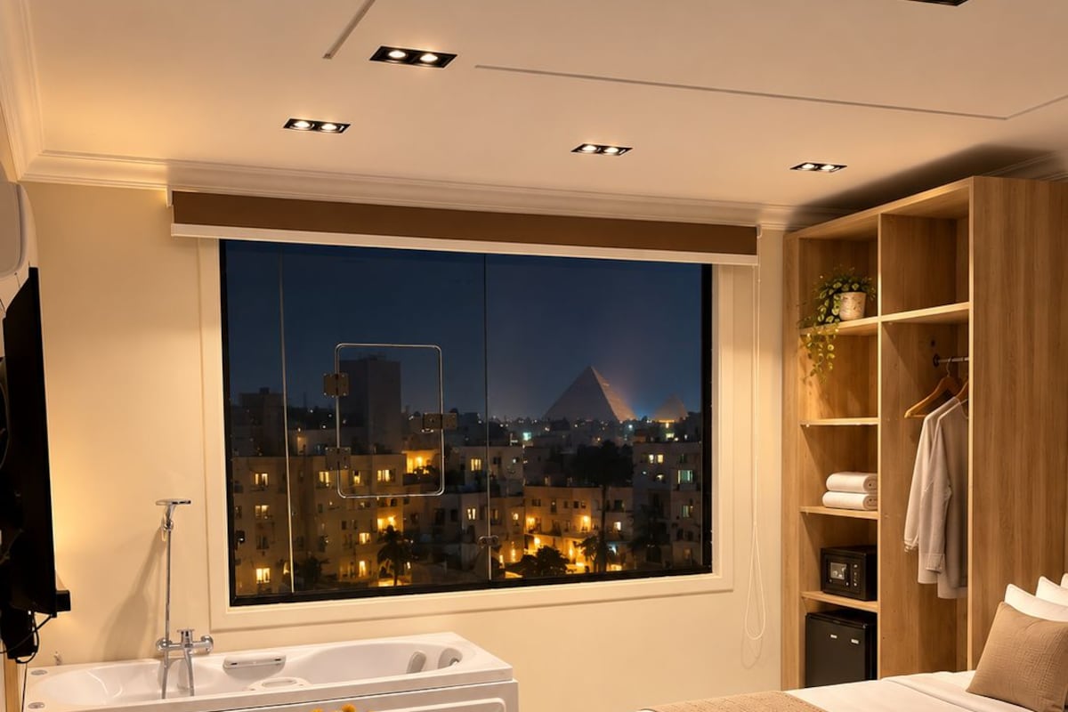 A modern bedroom with a large bed, a sitting area with a table and chair, a bathtub, and a view of a cityscape at night.