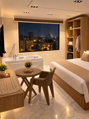 A modern bedroom with a large bed, a sitting area with a table and chair, a bathtub, and a view of a cityscape at night. - Wateen Pyramids View (Giza)