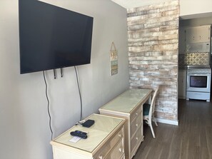 A room with a flat-screen TV mounted on the wall, a small table with a remote control, a wicker chair, and a kitchen area with a microwave and oven. - Hideaway Waterfront Resort & Hotel (Cape Coral)