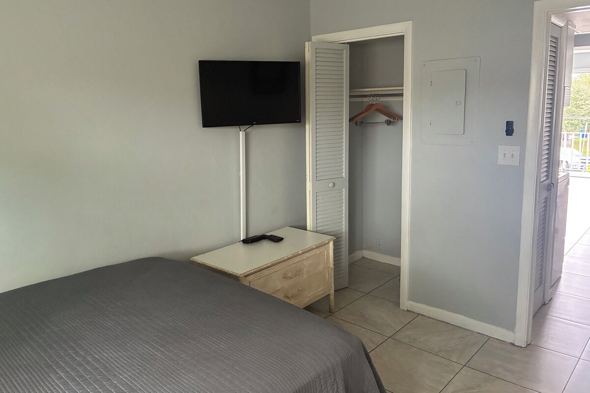 A bedroom with a bed, a bedside table, a TV mounted on the wall, a closet with a hanging rod, and an open door leading to a balcony.