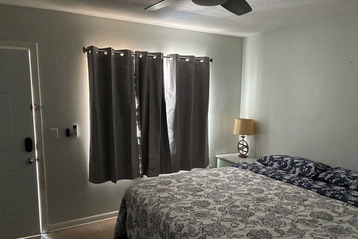 A bedroom with a bed, curtains, a nightstand with a lamp, and a door.