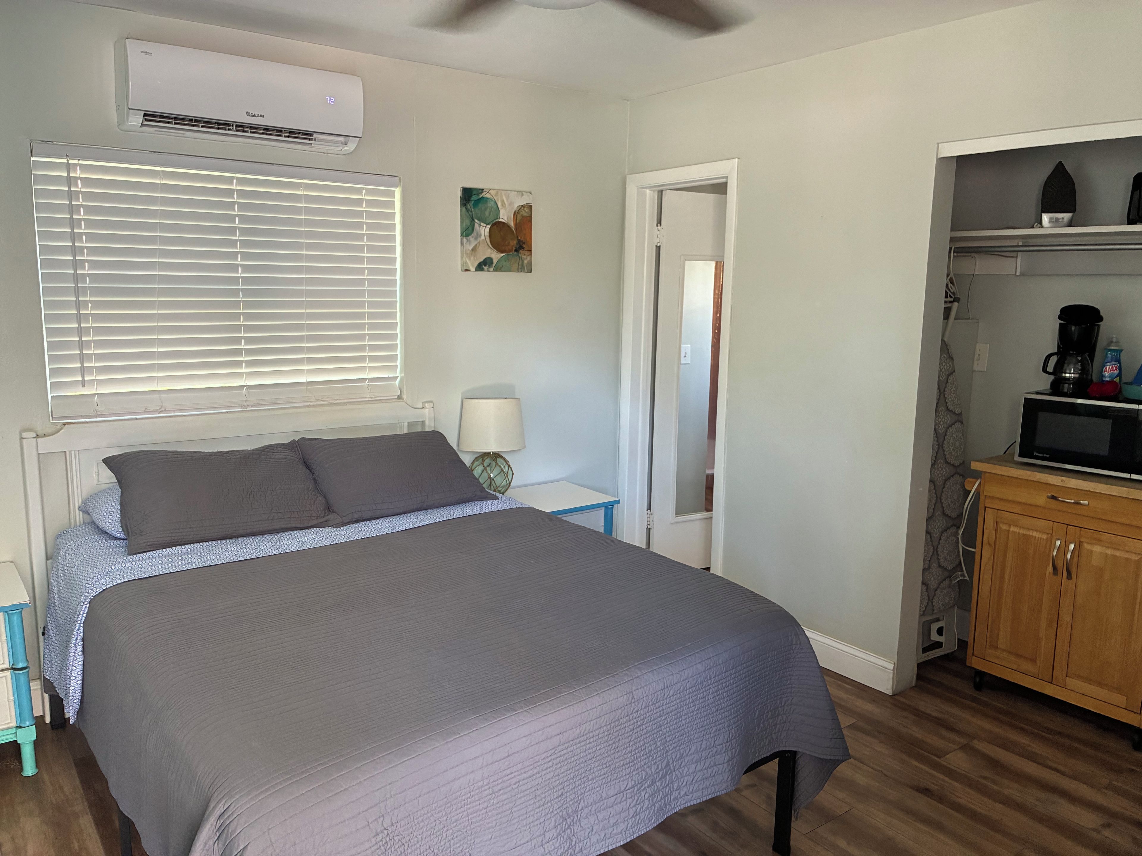 A bedroom with a bed, a nightstand, a lamp, a door, and a kitchenette area with a microwave.