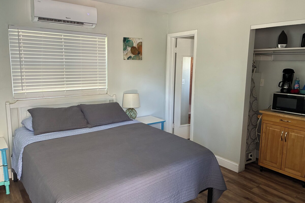 A bedroom with a bed, a nightstand, a lamp, a door, and a kitchenette area with a microwave.
