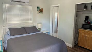 A bedroom with a bed, a nightstand, a lamp, a door, and a kitchenette area with a microwave.