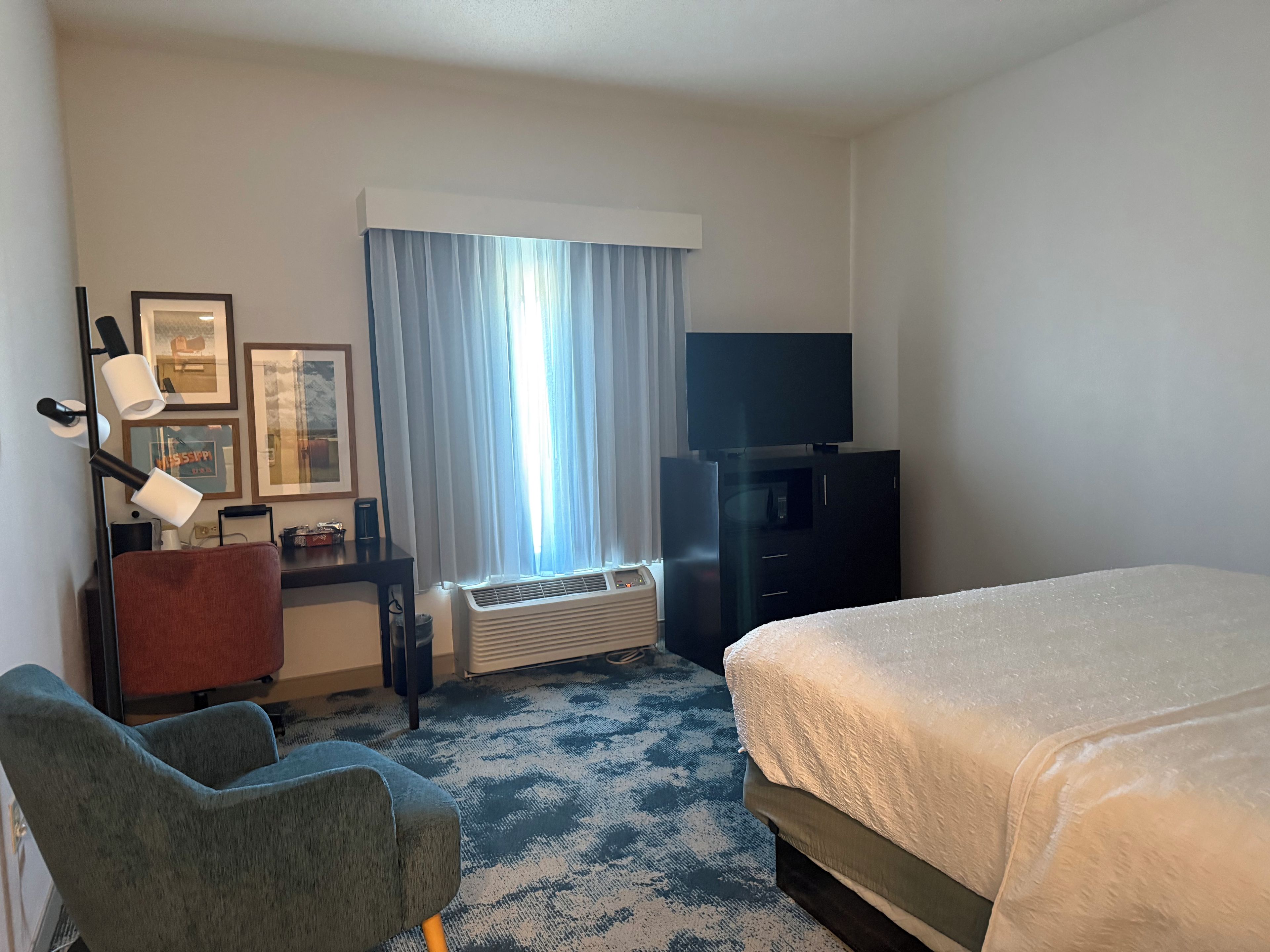 A hotel room with a bed, a desk with a chair, a television, a window with curtains, and a wall-mounted air conditioner.