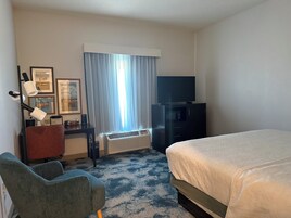 A hotel room with a bed, a desk with a chair, a television, a window with curtains, and a wall-mounted air conditioner.