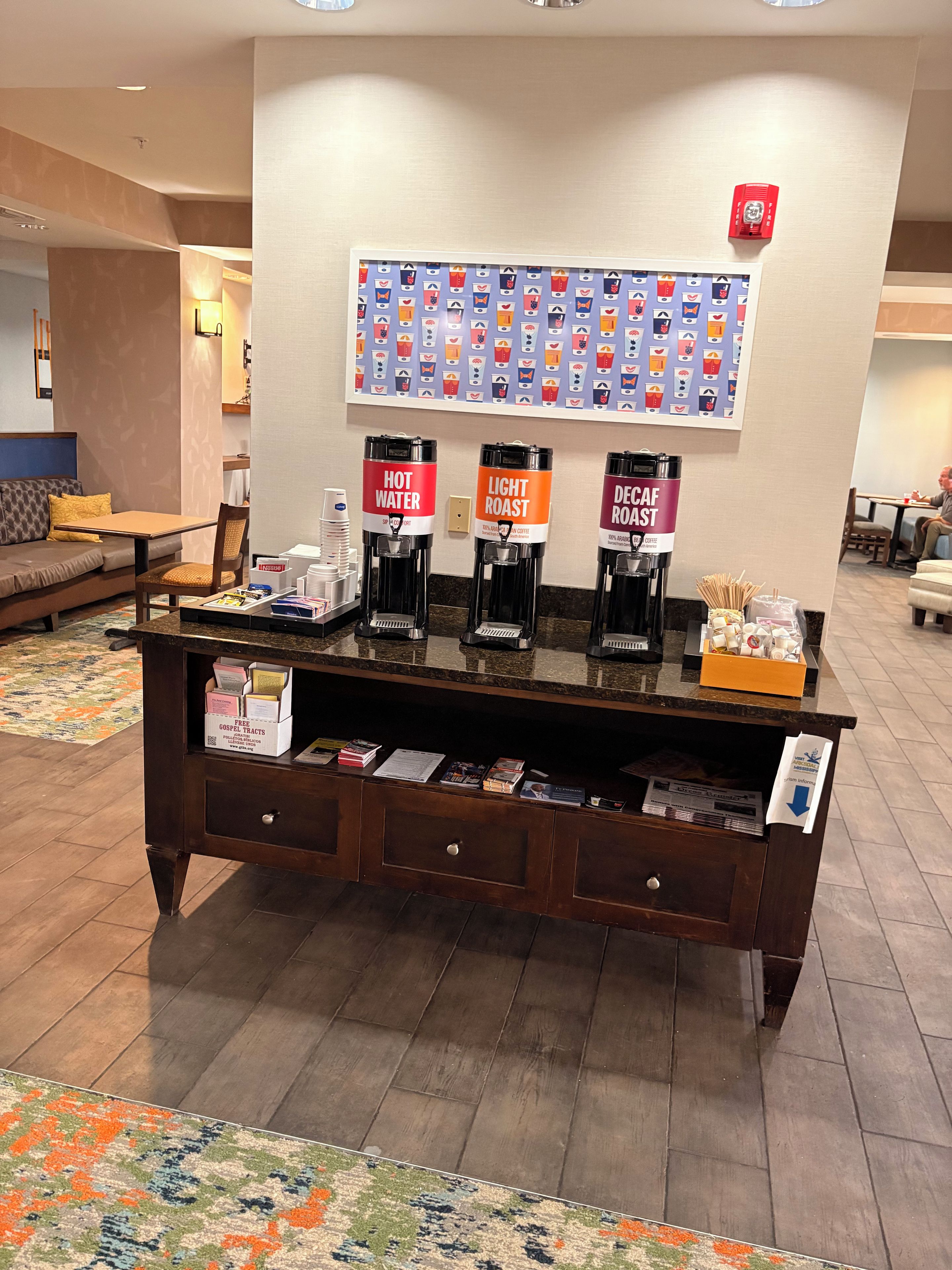 A hotel reception area with a coffee station, a wall with a colorful pattern, and a sofa in the background.