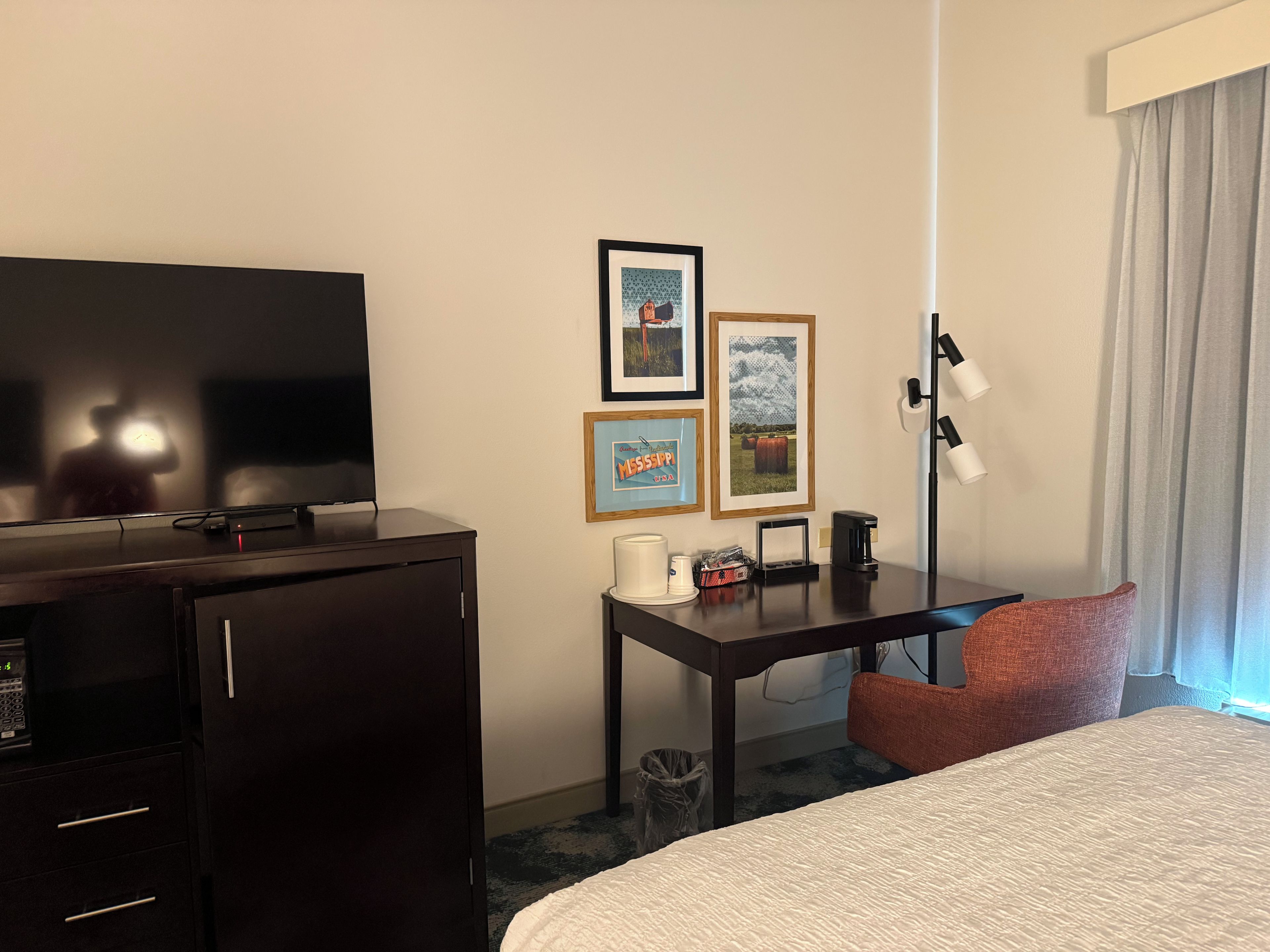 A hotel room with a bed, a desk with a lamp, a television, and framed pictures on the wall.
