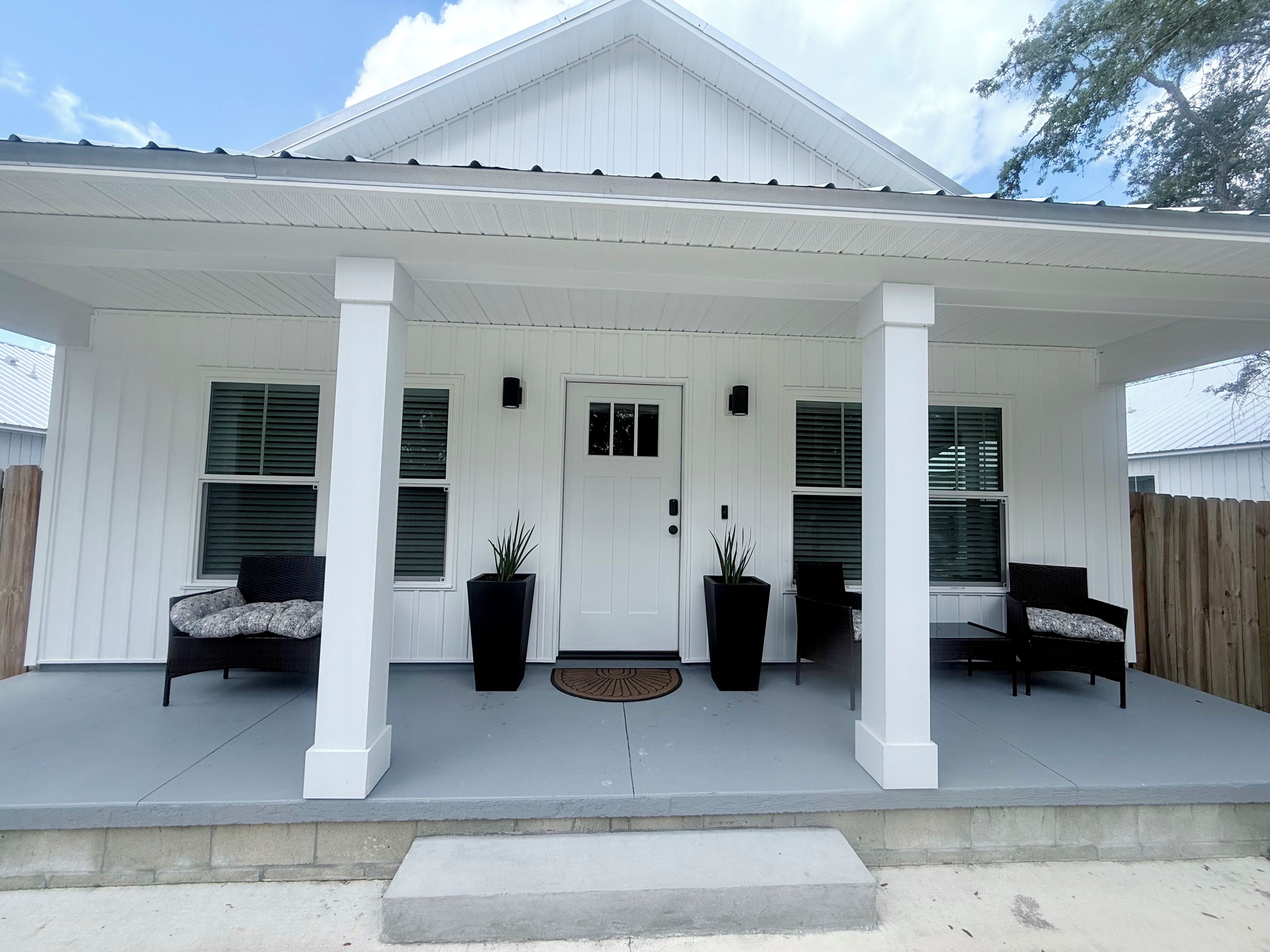 A house with a covered porch, two potted plants, a door, and two chairs.