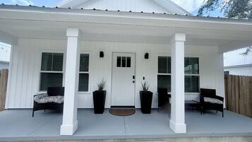A house with a covered porch, two potted plants, a door, and two chairs.