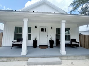 A house with a covered porch, two potted plants, a door, and two chairs.