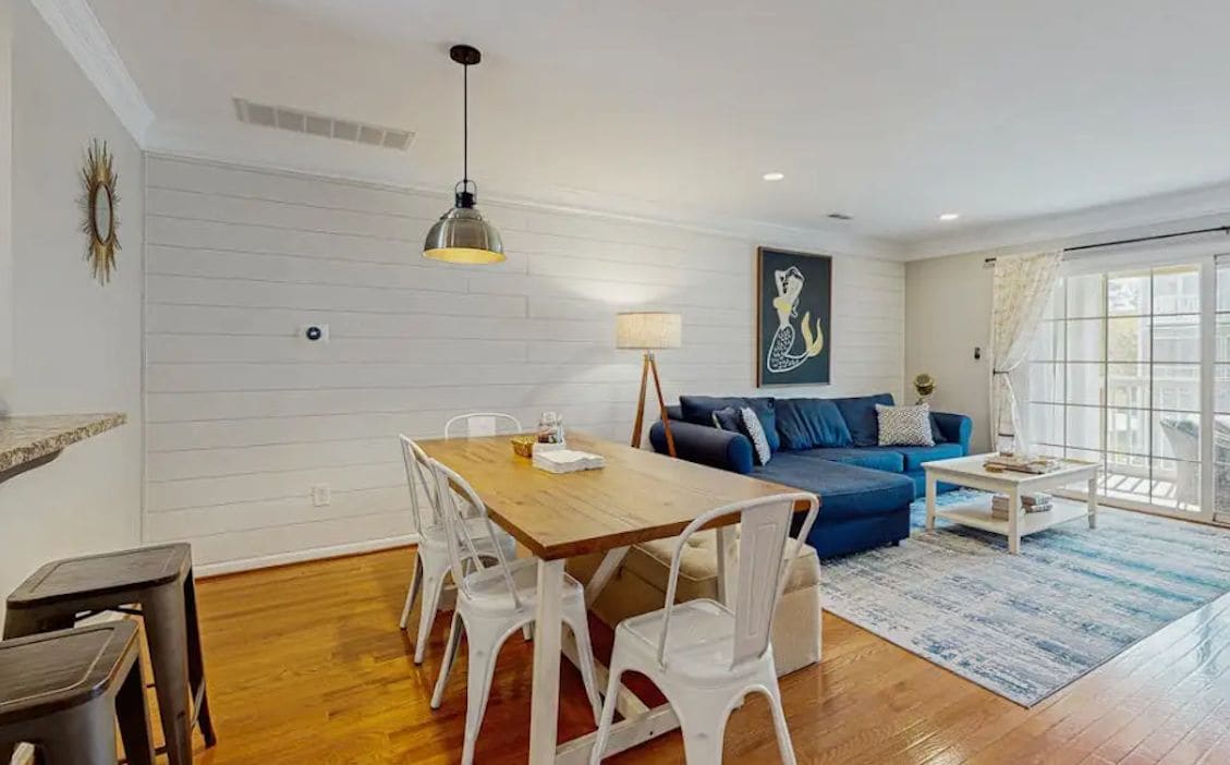 A modern living room with a wooden dining table, white chairs, a blue sofa, and a large window with curtains.