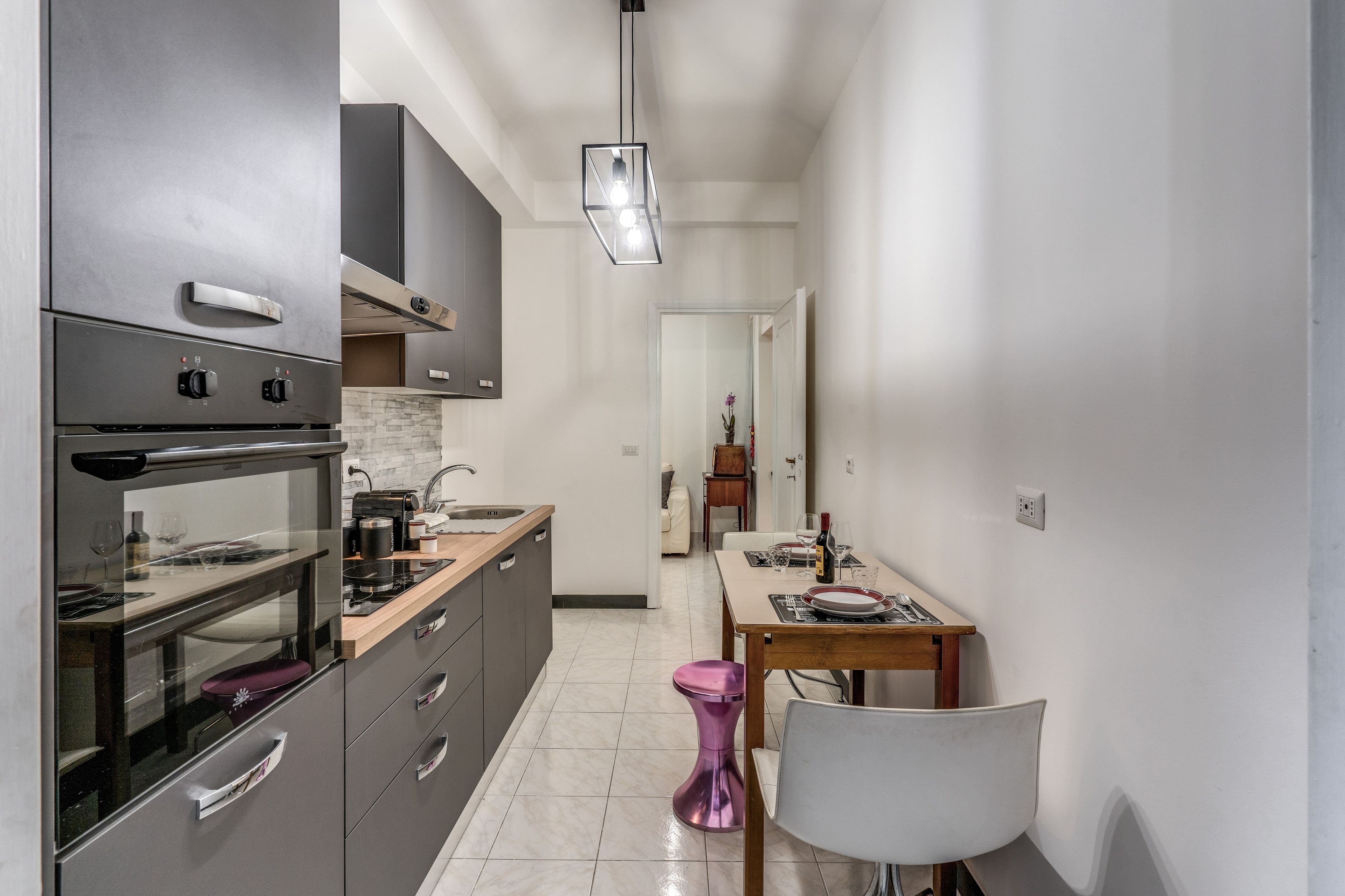 A modern kitchen with a built-in oven, a dining area with a table and chairs, and a hallway leading to a bedroom.