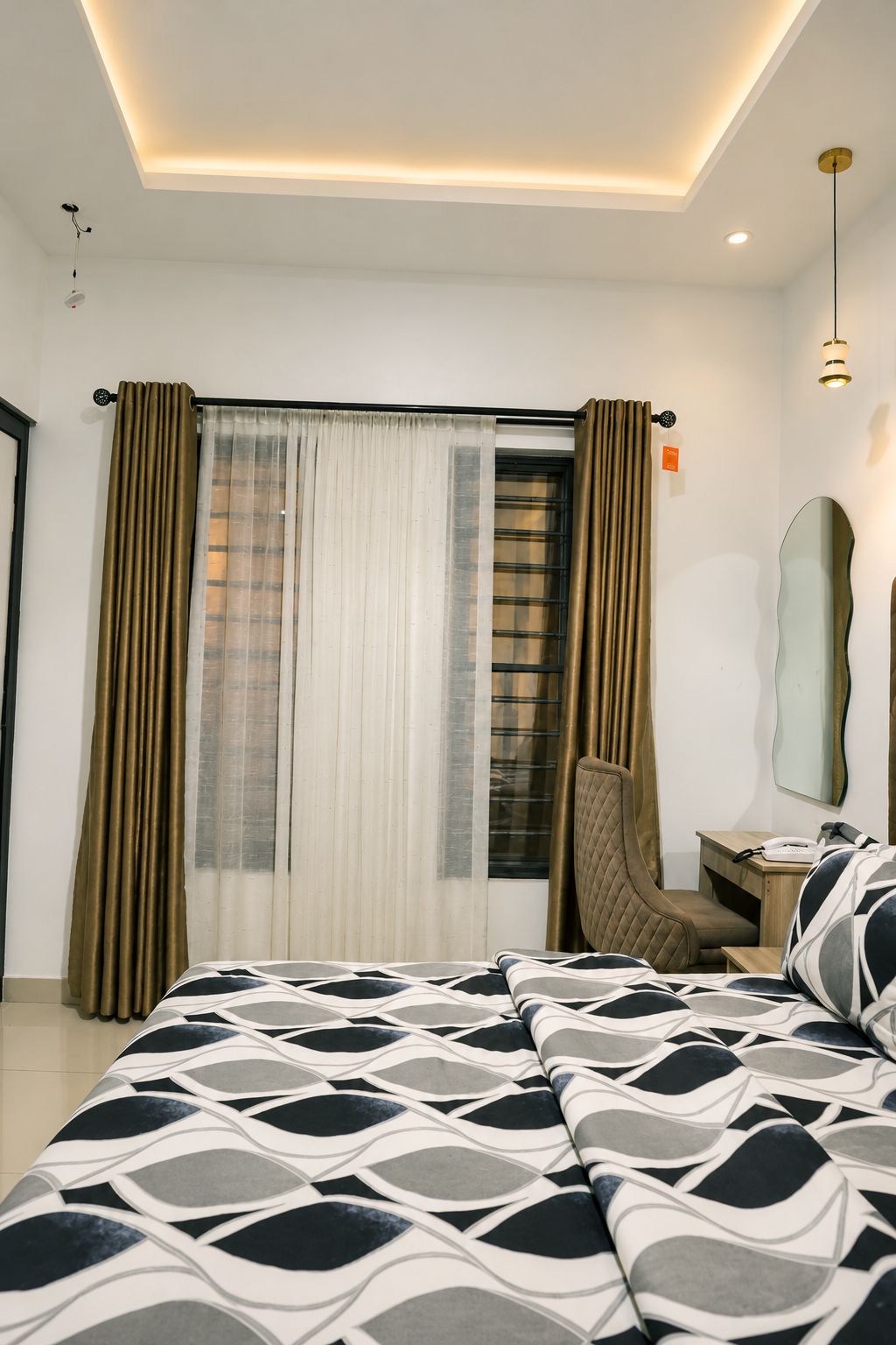 Une chambre d’hôtel avec un lit, un bureau, une chaise, un miroir et une fenêtre avec des rideaux.