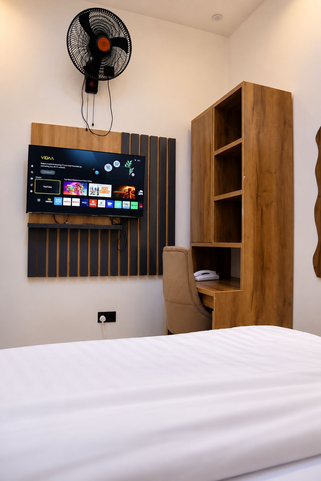 Une chambre d’hôtel moderne équipée d’un téléviseur à écran plat fixé au mur, qui affiche l’interface d’un service de diffusion en continu, d’une bibliothèque en bois, d’un bureau avec une chaise et d’un lit avec une literie blanche.