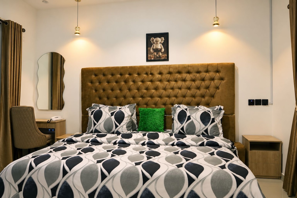 Quarto de hotel com cabeceira estofada, cama com roupa de cama estampada, travesseiro verde de destaque, mesa de cabeceira, cadeira, espelho e quadro pendurado na parede.